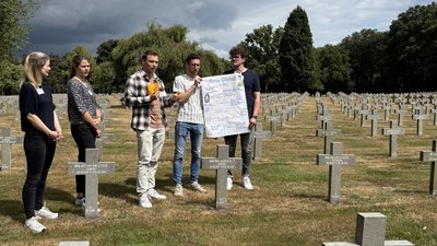 Zwei junge Frauen und drei junge Männer stehen auf einem Friedhof zwischen Grabkreuzen. Sie halten ein selbstgestaltetes Plakat.